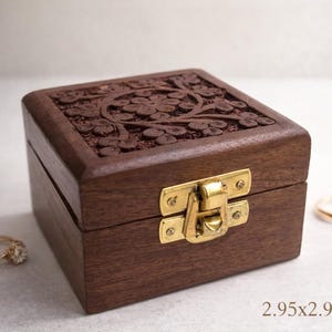 May include: A square, dark brown wooden box with a floral carved lid and a brass clasp. The box measures 2.95 x 2.95 x 1.97 inches. Two gold rings are visible near the box, suggesting it is a jewelry box.