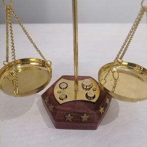 May include: A decorative brass scale with two round pans suspended by chains. The base is a dark wood hexagon with brass star accents. The scale's central support is a vertical brass rod.