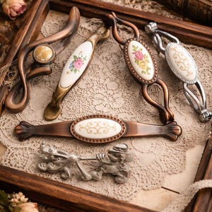 Peut inclure: Divers modèles de poignées et de tirettes de meubles de style vintage. Les pièces présentent des motifs floraux et des finitions métalliques, comme le bronze et l'argent. Elles sont disposées sur une nappe en dentelle, mettant en valeur les détails.