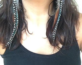 May include: Long feather earrings with a mix of colors, including black, white, teal, and brown. The earrings are hanging from a black string and are worn by someone wearing a black tank top.