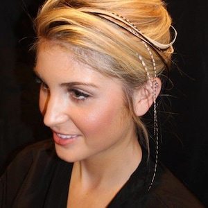 May include: A woman with blonde hair is wearing a black feather hair extension. The extension is attached to her hair with a small clip. The feather is long and flowing, and it is a light brown color.