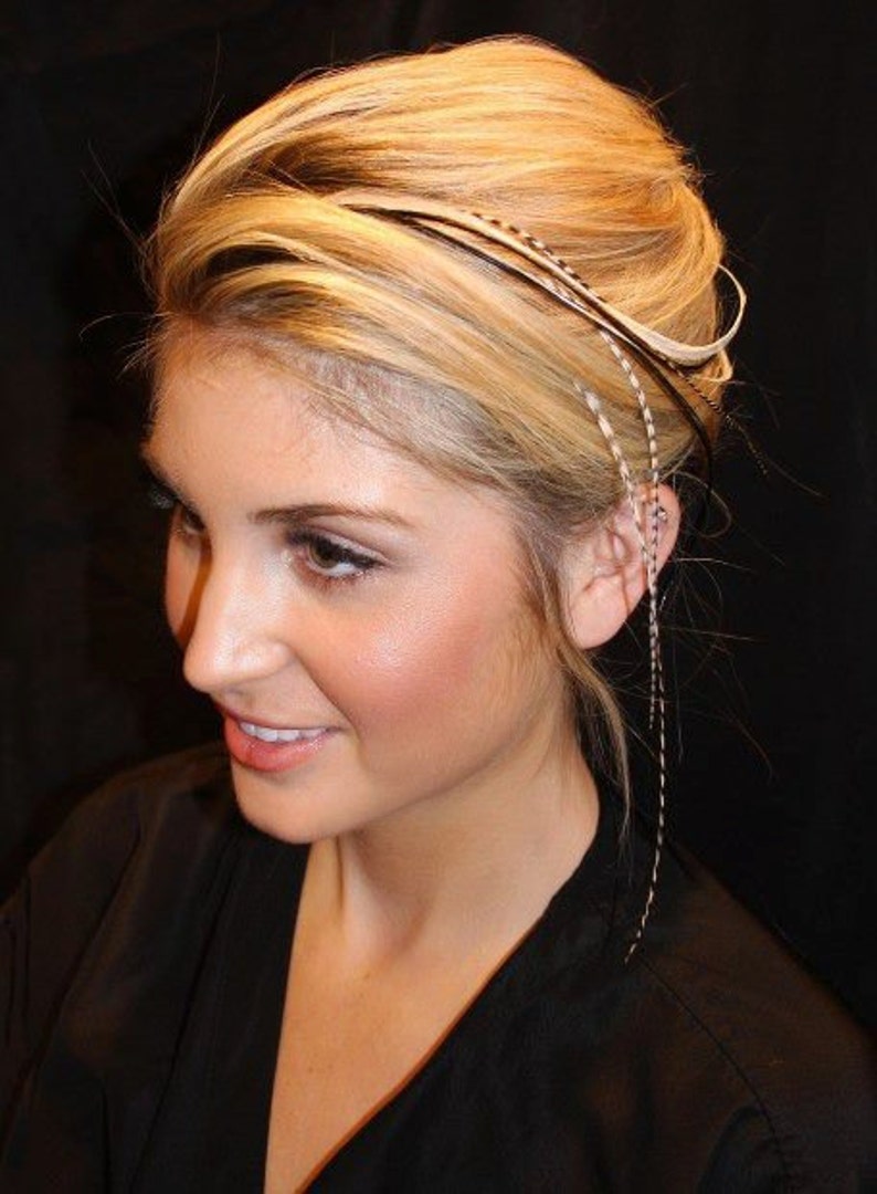 May include: A woman with blonde hair is wearing a black feather hair extension. The extension is attached to her hair with a small clip. The feather is long and flowing, and it is a light brown color.