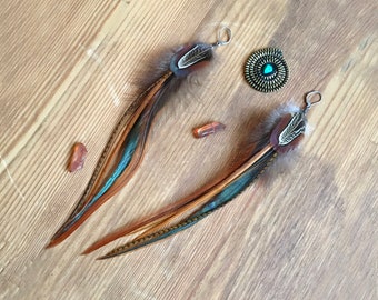 May include: A pair of long feather earrings with brown, blue, and white feathers. The earrings have a silver hook and a small brown bead at the top. The earrings are on a wooden surface.