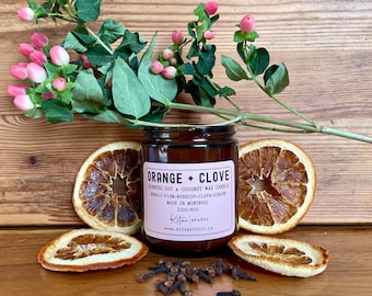 May include: A brown glass jar candle with a pink label that reads "ORANGE + CLOVE". The candle is surrounded by dried orange slices, cloves, and a sprig of greenery with pink berries. The candle is made in Montreal.