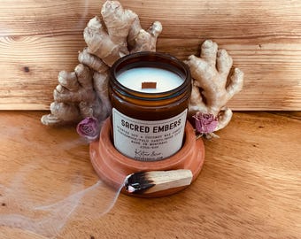 May include: A brown glass jar candle with a white label that reads "SACRED EMBERS". The candle sits on a terracotta-colored holder, with a lit palo santo stick and ginger root in the background. A small pink rose is also visible.
