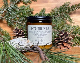 May include: A brown glass candle jar with a black lid. The label reads "INTO THE WILD" and lists the scent as "SCENTED SOY & COCONUT WAX CANDLE PALO SANTO-SAGE-CEDARWOOD-CYPRESS CLEAR-CLEANSE-GROUND MADE IN MONTREAL 2300/60H Kstper &co ESTAANDCO.COM".