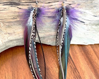 May include: A pair of long feather earrings with purple, black, white, and teal feathers. The earrings are hanging from a wooden surface.