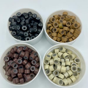 Peut inclure: Quatre petits bols blancs remplis de micro-anneaux pour extensions de cheveux. Les bols contiennent des micro-anneaux noirs, bruns, brun clair et blonds.