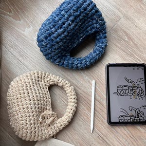 May include: A top-down shot features two handmade crochet items, one blue and one beige, each with a handle. A tablet displays a crochet pattern, and a white stylus lies nearby. The items are on a wooden surface.