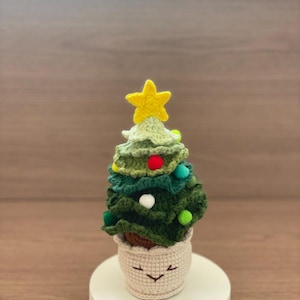 May include: A crocheted Christmas tree in a smiling pot. The tree is made of green yarn and topped with a yellow star. Colorful pom-poms are used as ornaments. The pot is beige with a smiling face.