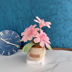 以下が含まれることがあります： ピンクの花と緑の葉が特徴の手作りのかぎ針編みのフラワーアレンジメント。ベージュの鉢に入っており、「Hand Made」とスマイリーフェイスのタグが付いています。白い台座の上に置かれ、背景には青い時計があります。
