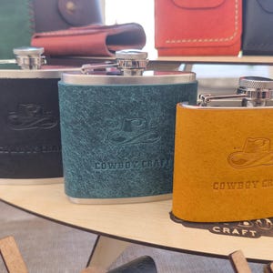 Può includere: Tre fiaschette in acciaio inossidabile rivestite in pelle testurizzata nera, verde ottanio e giallo senape. Ogni fiaschetta presenta un'icona di cappello da cowboy in rilievo e la scritta COWBOY CRAFT. I tappi in metallo sono argentati e zigrinati.