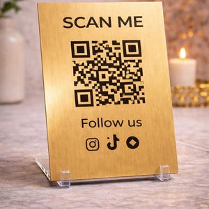 May include: A gold-coloured sign displaying "SCAN ME" and a QR code. Beneath the code, the sign reads "Follow us" with social media icons. The sign is supported by clear acrylic stands.