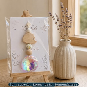 Könnte beinhalten: Ein Sonnenfänger mit einem Holz-Igel, einer Blumenperle, einer schlichten Holzperle und einer facettierten, irisierenden Kristallkugel, befestigt an einer Karte mit der Aufschrift 'Zu Taufe' und '16. August 2026', präsentiert auf einer kleinen Holzstaffelei.