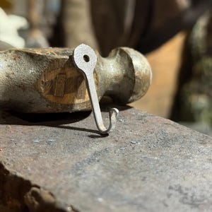 Puede incluir: Un pequeño gancho de metal forjado a mano con una sección superior plana y redondeada que presenta un orificio circular preperforado. El gancho tiene un extremo inferior curvo en forma de J y un acabado metálico gris mate con textura.
