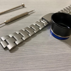 May include: A selection of watch repair tools and components. The image shows a silver metal watch strap, two small screwdrivers, and a black and blue loupe. The tools are arranged on a light grey surface, suggesting watch maintenance or repair.