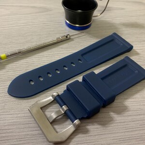 May include: A navy blue watch strap with a silver buckle, a small screwdriver, and a black and silver tool with a magnifying glass. The watch strap has several holes for adjustment.