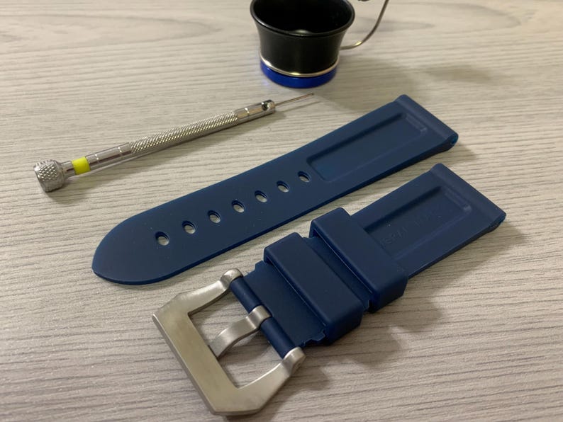 May include: A navy blue watch strap with a silver buckle, a small screwdriver, and a black and silver tool with a magnifying glass. The watch strap has several holes for adjustment.