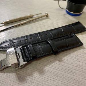 May include: Black leather watch strap with a crocodile pattern, featuring a silver clasp with the IWC logo. Two small watch repair tools and a magnifying glass are also present on a light-coloured surface.