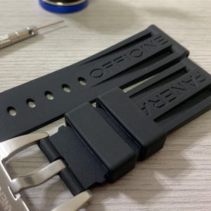 May include: Black watch strap with the brand name "PANERAI" and "OFFICINE" embossed. The strap is made of a flexible material and has a silver buckle. A small tool and a blue and gold object are in the background.