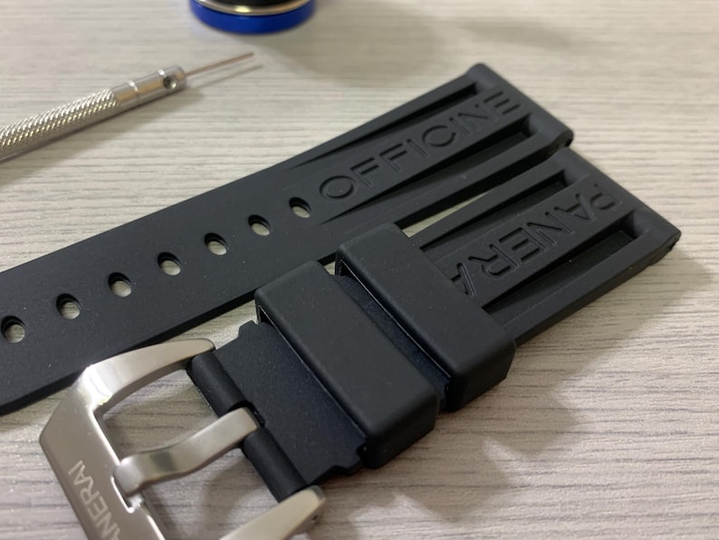 May include: Black watch strap with the brand name "PANERAI" and "OFFICINE" embossed. The strap is made of a flexible material and has a silver buckle. A small tool and a blue and gold object are in the background.