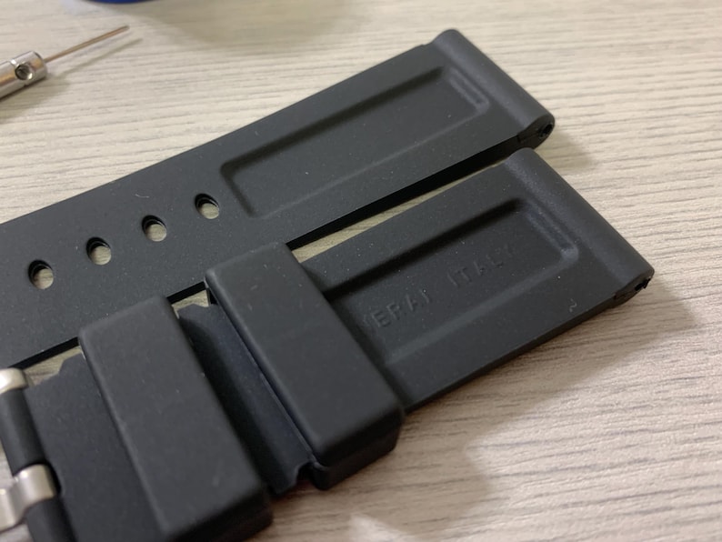 May include: Close-up of two black watch straps with a matte finish. One strap is partially connected to a silver clasp. The text "PANERAI ITALY" is embossed on one strap. A small tool is visible in the background.
