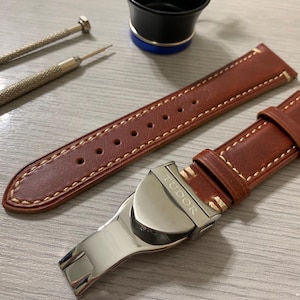 May include: A brown leather watch strap with cream stitching and a silver clasp, featuring the brand name "TUDOR". Two small screwdrivers and a black and blue object are also visible.