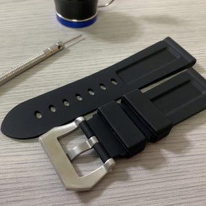 May include: Black rubber watch strap with a silver buckle. The strap has several holes for adjustment. A small silver tool with a yellow handle is next to the strap. A black and blue object is in the background.