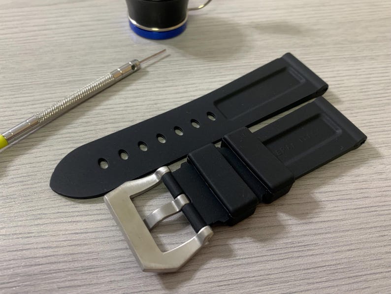 May include: Black rubber watch strap with a silver buckle. The strap has several holes for adjustment. A small silver tool with a yellow handle is next to the strap. A black and blue object is in the background.