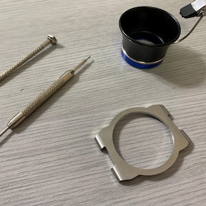 May include: A selection of watch repair tools, including two small screwdrivers, a black and blue loupe, and a silver watch case holder. The tools are arranged on a light grey surface.