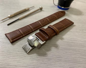 May include: Brown leather watch strap with a crocodile pattern and a silver clasp. Two small watch repair tools are also present. The watch band is a rich brown color and appears to be new.