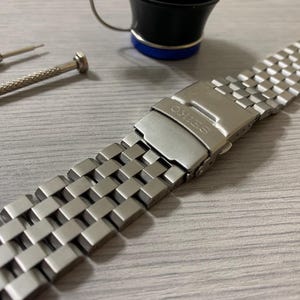 May include: A stainless steel watch band with a brushed finish, featuring a Seiko clasp. The band is composed of square links and lies on a light gray surface. Two small screwdrivers and a small black cup are also visible.
