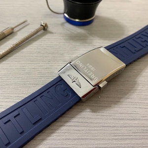 Puede incluir: Una correa de reloj Breitling azul marino con un cierre plateado. La correa está hecha de un material flexible y presenta el nombre de la marca en relieve. En el fondo se ven dos pequeñas herramientas de reparación de relojes.