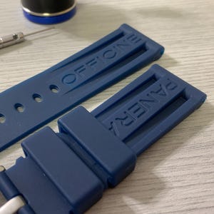 May include: Close-up of a navy blue watch strap with the brand name "OFFICINE PANERAI" embossed on the rubber. A small screwdriver and a watch component are visible in the background.