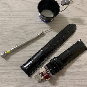 May include: Black leather watch straps with a crocodile pattern, a watchmaker's loupe, and a small screwdriver. The watch straps have a silver clasp with the brand name. The items are on a light wood grain surface.