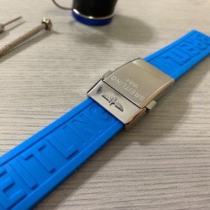 May include: A bright blue watch strap with a silver clasp. The strap is made of a flexible material and features the brand name "BREITLING 1884" on the clasp and the brand name embossed on the strap. Two small screwdrivers are visible.