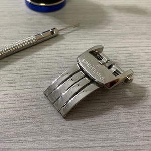 May include: A silver Breitling watch clasp with a polished finish, featuring the brand name. A small silver tool and a blue and silver object are also visible. The clasp is on a light gray wooden surface.