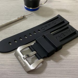 May include: Black watch strap with the word "OFFICINE" and "PANERAI" embossed. The strap has a silver buckle with the brand name engraved. A small tool and a black and blue object are also in the image.