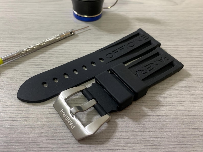 May include: Black watch strap with the word "OFFICINE" and "PANERAI" embossed. The strap has a silver buckle with the brand name engraved. A small tool and a black and blue object are also in the image.