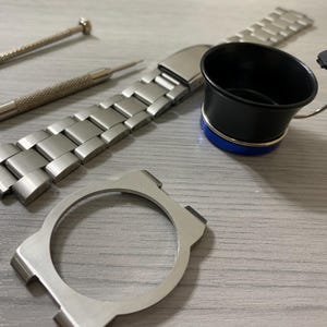 May include: A collection of watch repair tools and components, including a silver watch band, a watch case, a black and blue loupe, and two small screwdrivers. The items are arranged on a light gray surface.
