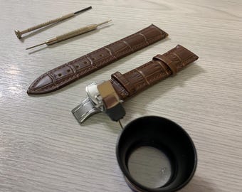 May include: A brown leather watch strap with a crocodile pattern, a silver clasp, and two small screwdrivers. A magnifying glass is also present, suggesting watch repair or maintenance.