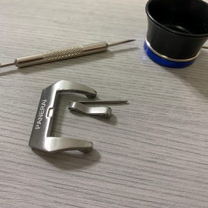 Puede incluir: Una hebilla de reloj de acero inoxidable con el nombre de la marca "Panerai" grabado, junto con una barra de resorte y una herramienta de reparación de relojes. También hay una lupa negra y azul.