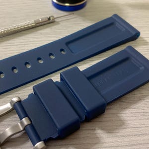 May include: A navy blue watch strap with a stainless steel buckle and a matching blue strap. The strap has several holes for adjustment and the text "ERA ITALY" is visible. A small tool and a blue cap are also in the image.