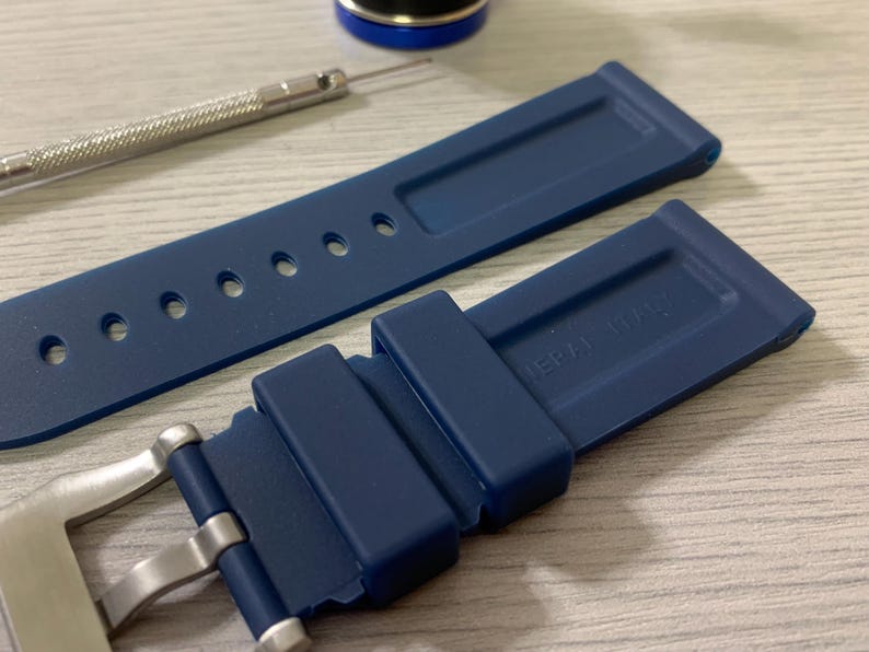 May include: A navy blue watch strap with a stainless steel buckle and a matching blue strap. The strap has several holes for adjustment and the text "ERA ITALY" is visible. A small tool and a blue cap are also in the image.