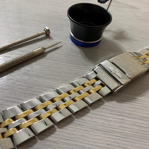 Könnte beinhalten: Ein Edelstahl- und goldfarbenes Uhrenarmband mit Faltschließe. Zwei Uhrenreparaturwerkzeuge und eine schwarze Lupe mit blauem Sockel sind ebenfalls vorhanden. Das Armband ist etwa 18 cm lang.