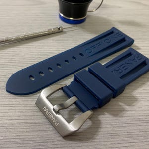 May include: A navy blue watch strap with a silver buckle, featuring the brand name "PANERAI". The strap is made of a flexible material and has the word "OFFICINE" embossed on the strap. A small screwdriver and a black and silver cup are also in the image.