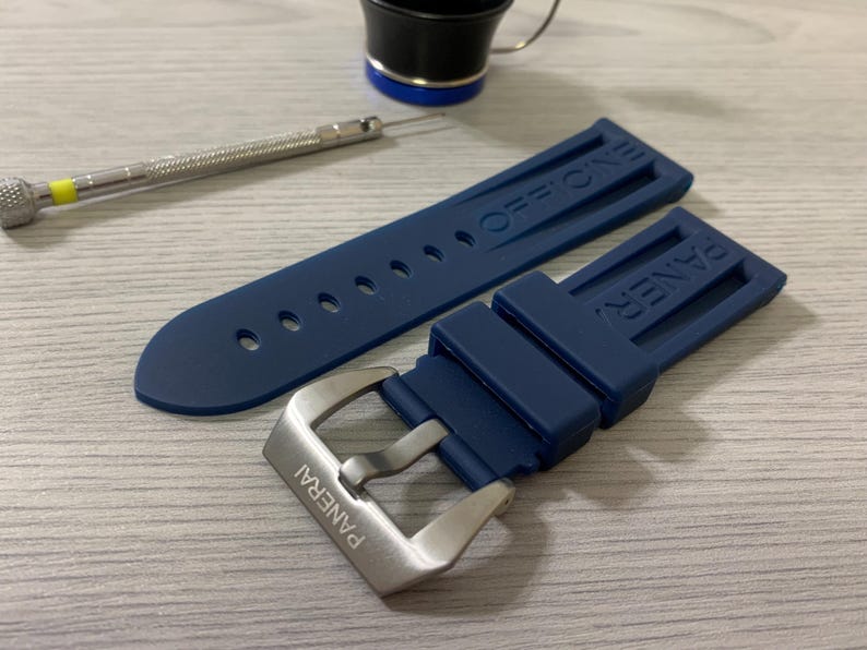 May include: A navy blue watch strap with a silver buckle, featuring the brand name "PANERAI". The strap is made of a flexible material and has the word "OFFICINE" embossed on the strap. A small screwdriver and a black and silver cup are also in the image.
