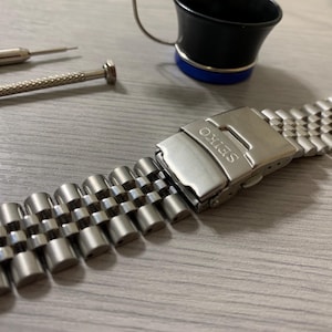 May include: Close-up of a stainless steel watch band with a SEIKO clasp. Two small screwdrivers and a black and blue loupe are in the background. The watch band is made of small, connected, rounded metal links.