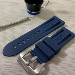 May include: A navy blue watch strap with the word "OFFICINE" embossed on one section and "PANERAI" on the other. A silver buckle is attached. A small screwdriver and a black and blue tool are also in the image.
