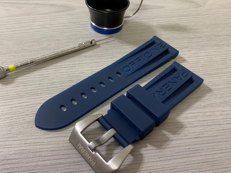 May include: A navy blue watch strap with the word "OFFICINE" embossed on one section and "PANERAI" on the other. A silver buckle is attached. A small screwdriver and a black and blue tool are also in the image.